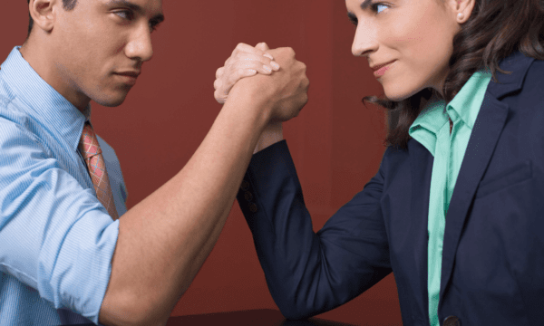 Medium shot of a man (left, wearing a blue button-down) and woman (right, wearing a mint green button-down and navy blazer) arm wrestling