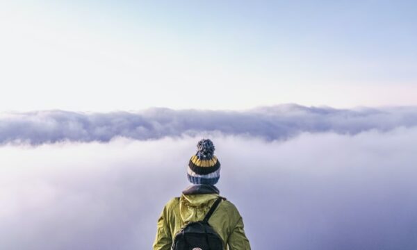 A hiker wearing a backpack and hat staring at the horizon, obscured by clouds