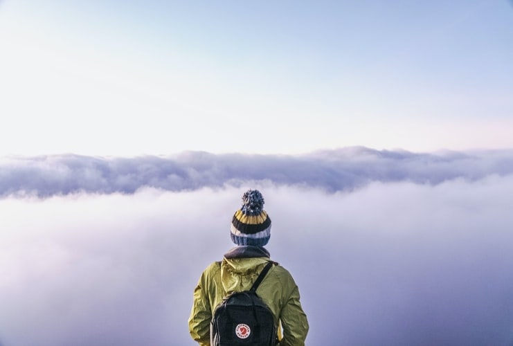 A hiker wearing a backpack and hat staring at the horizon, obscured by clouds