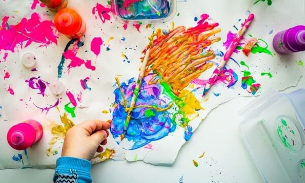 Blue, orange, pink, and yellow paint splattered across a white background, with a hand reaching for a paintbrush