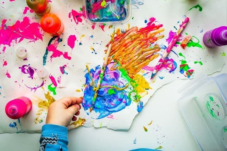 Blue, orange, pink, and yellow paint splattered across a white background, with a hand reaching for a paintbrush