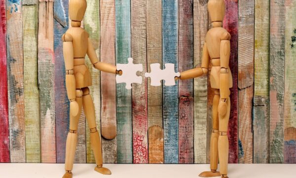 Photo of two wooden art model dummies facing each other, each holding a white puzzle piece, as if they're going to fit them together. Background is a wooden fence painted in a muted rainbow of colors.