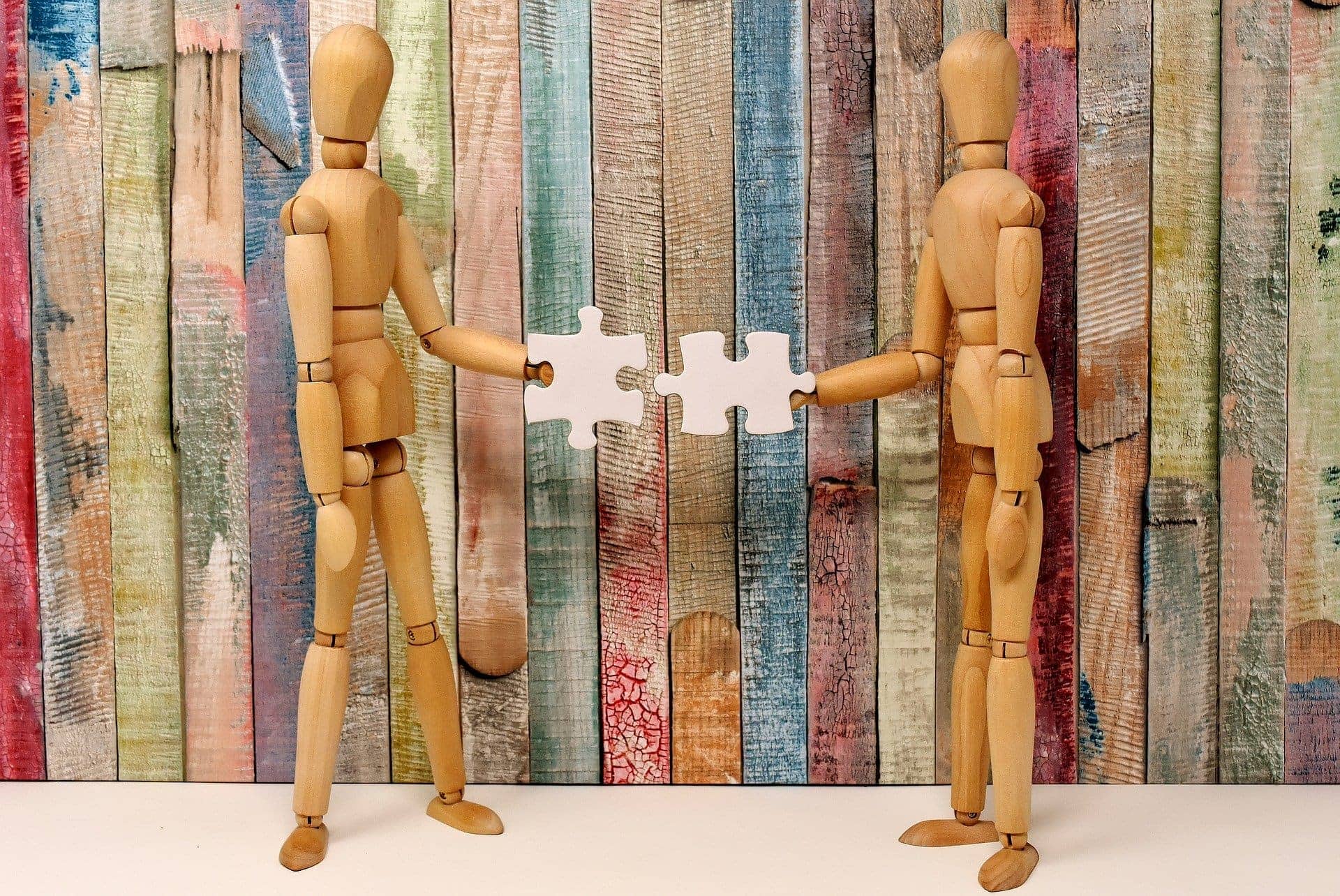 Photo of two wooden art model dummies facing each other, each holding a white puzzle piece, as if they're going to fit them together. Background is a wooden fence painted in a muted rainbow of colors.