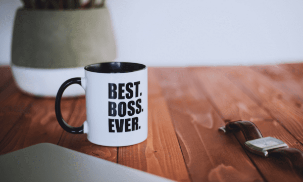 Photo of a dark polished wood floor with mug in the foreground (white, with black interior and black handle) that reads "Best. Boss. Ever." in all-caps black block letters. Potted plant and plain white wall in background.