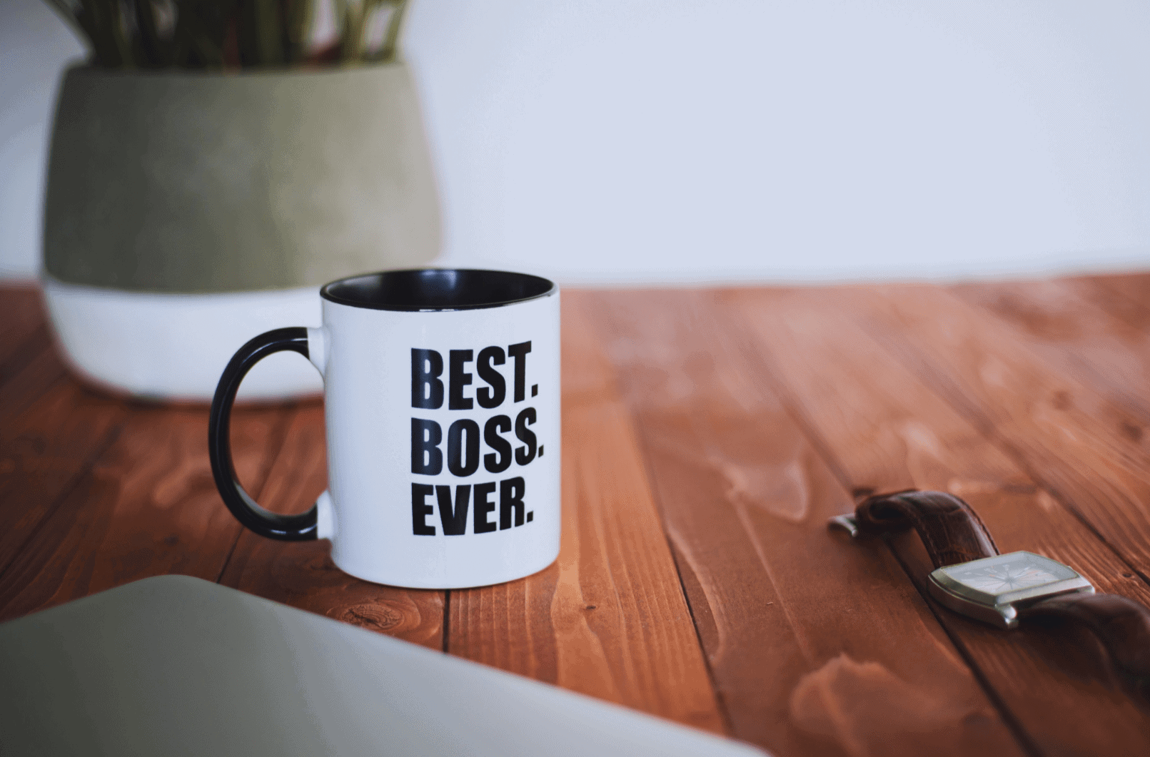 Photo of a dark polished wood floor with mug in the foreground (white, with black interior and black handle) that reads "Best. Boss. Ever." in all-caps black block letters. Potted plant and plain white wall in background.