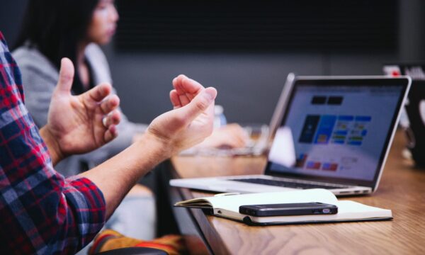 A pair of gesturing hands in the foreground (person is seated at a table on which an open laptop is in soft focus in the background).